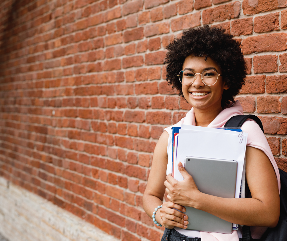 Theological Education Trends, A young black female student holds books in front of a red brick wall while smiling