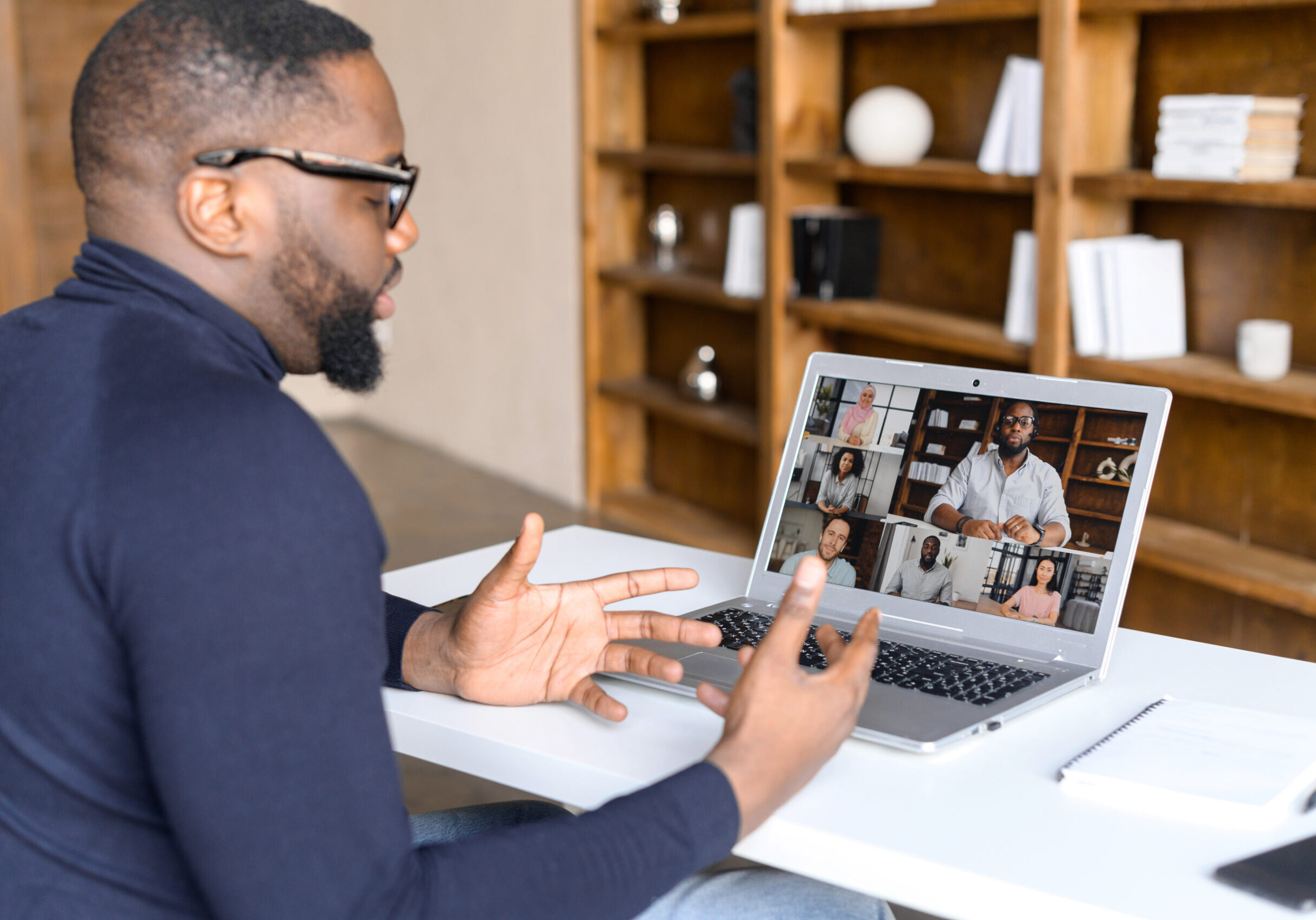 Virtual conference with employees, online briefing. Side view of a young African man in casual wear using laptop for video call, has video meeting with several people together. Remote work concept