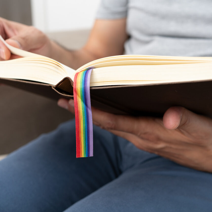 Happy man reading a paper book with LGTB flag sitting on a sofa at home